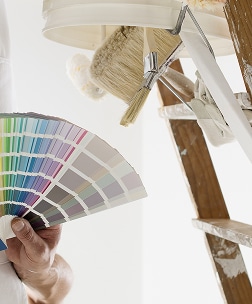 Choix de Couleurs Peinture : Nuancier et Outils Pro Peintre examinant un éventail de nuancier de couleurs, avec des pinceaux et une échelle en arrière-plan pour la décoration intérieure.