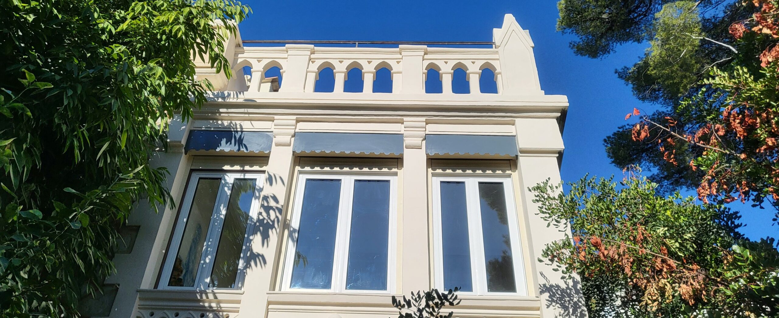 Architecture de villa néo-gothique et façade élégante Façade de villa crème avec balustrade crénelée, fenêtres blanches et stores gris sous un ciel bleu azur.