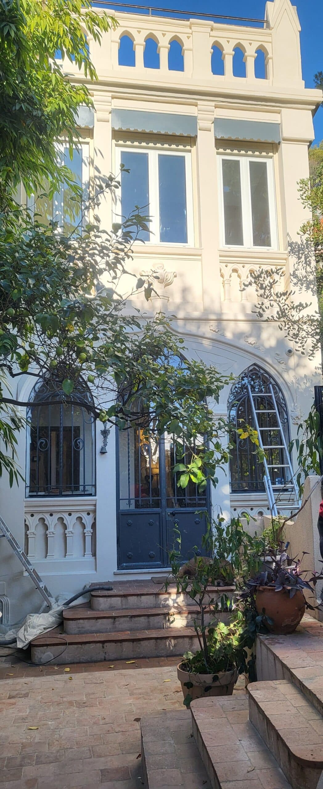 Façade d'une maison néo-gothique élégante avec fenêtres cintrées, plantes en pot et échelle de rénovation.