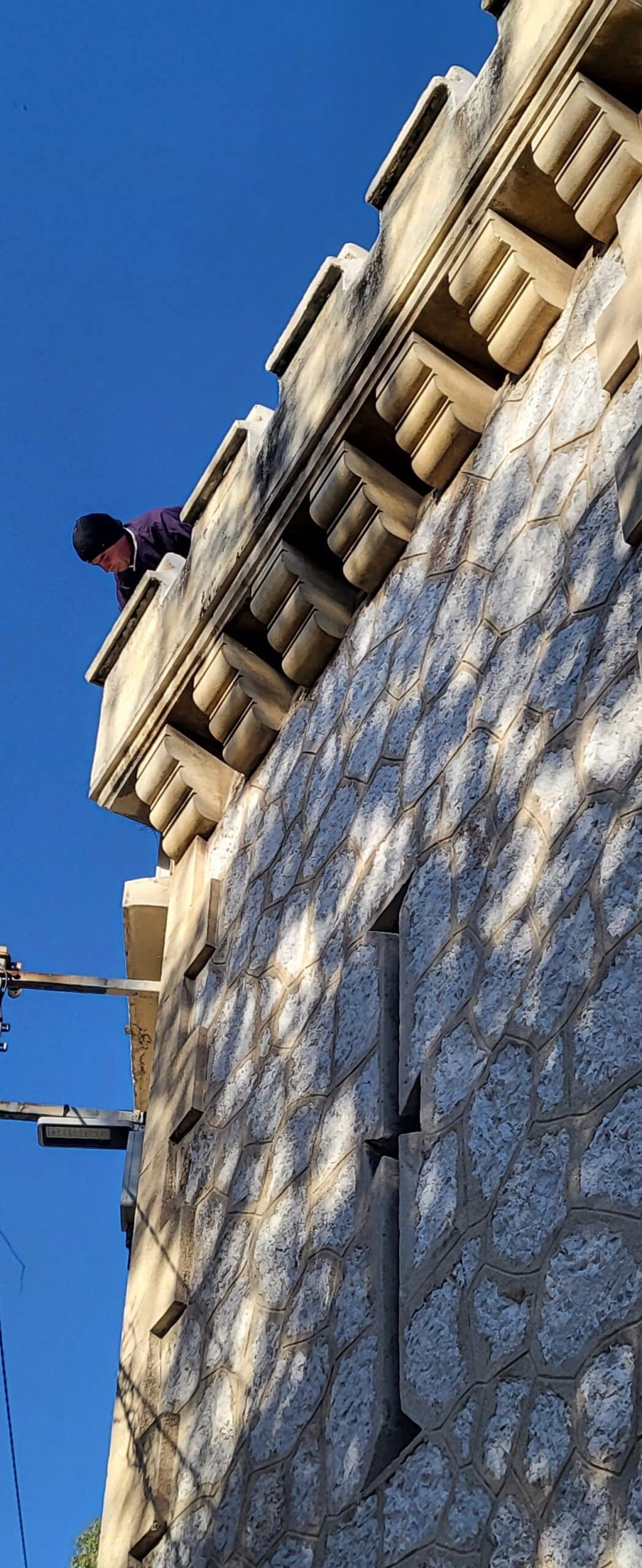 Homme observant du haut d'un bâtiment en pierre aux corniches sculptées sous un ciel bleu dégagé.