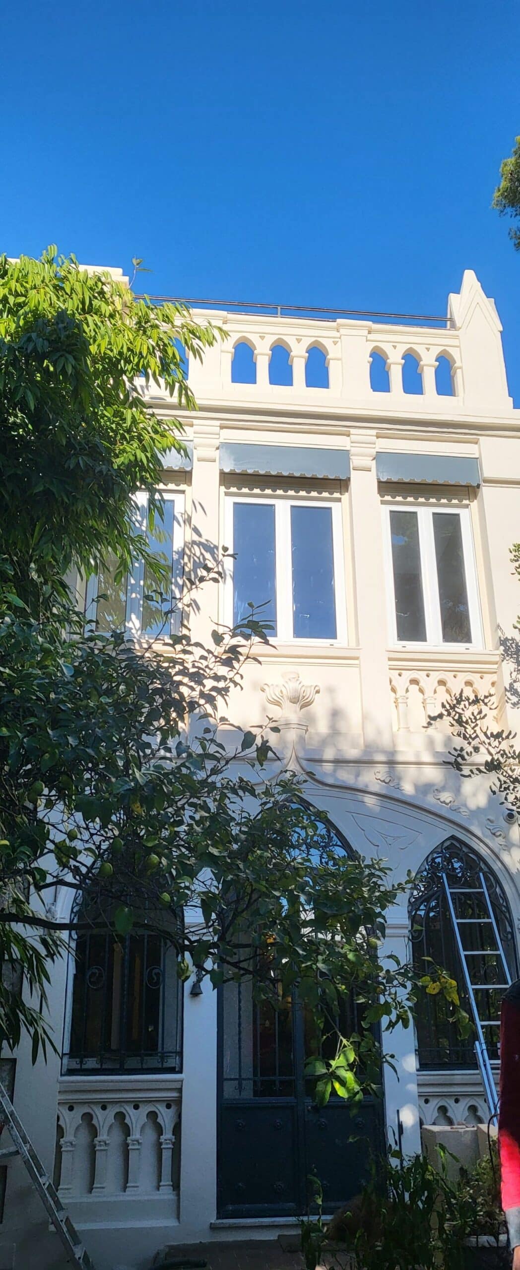 Maison blanche néogothique aux fenêtres en ogive bordée d'arbres verdoyants sous un ciel bleu éclatant.