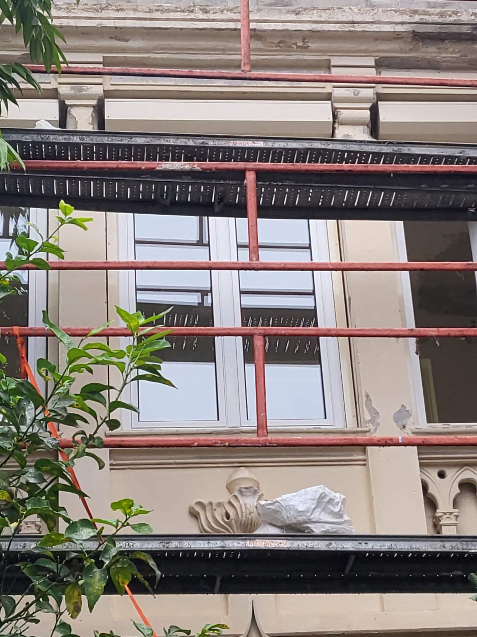 Échafaudage rouge devant une fenêtre de bâtiment ancien en rénovation avec détails architecturaux sculptés.