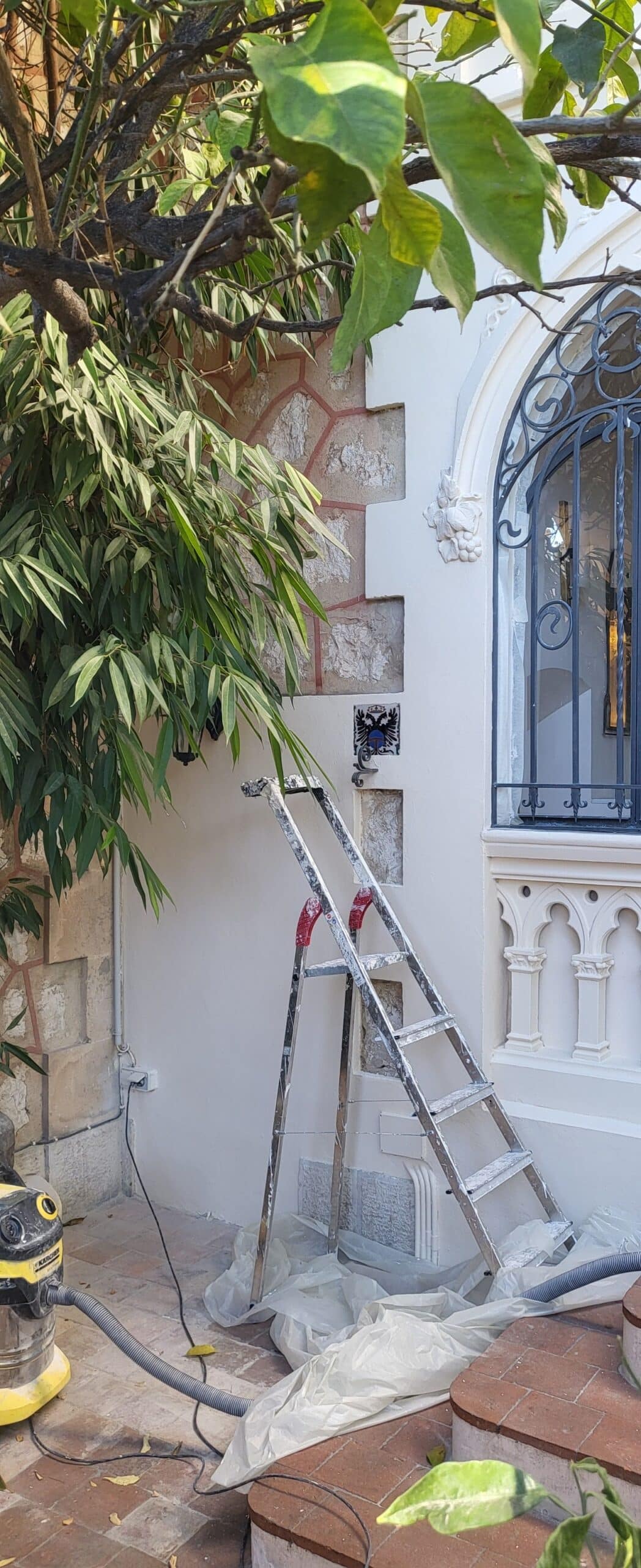 Escabeau et aspirateur Karcher devant une façade blanche en rénovation avec ornements gothiques et feuillage vert.