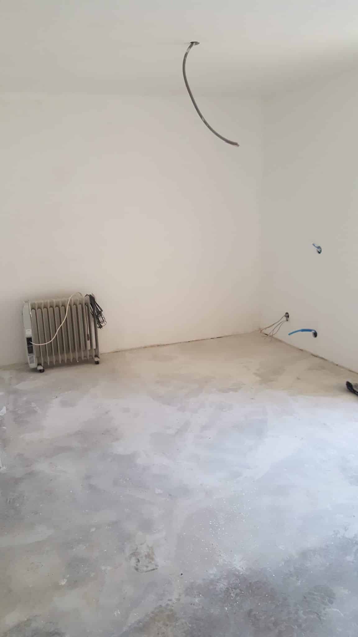 Pièce vide en rénovation avec murs blancs, sol en béton, radiateur d'appoint et câbles électriques apparents.