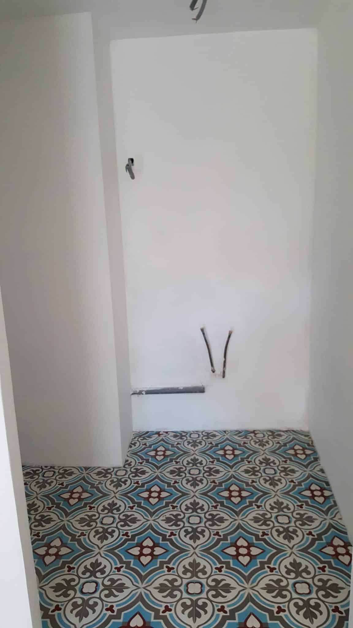 Coin salle de bain en travaux avec carrelage à motifs bleus et tuyaux de plomberie sur mur blanc.